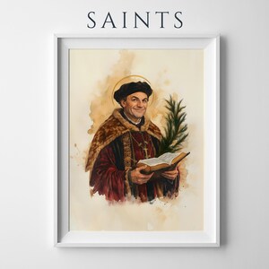 May include: A framed art print titled SAINTS featuring a watercolour-style portrait of a man in a dark cap and red robes with a fur collar, holding an open book and a green palm frond. A thin gold halo circles his head against a beige background.