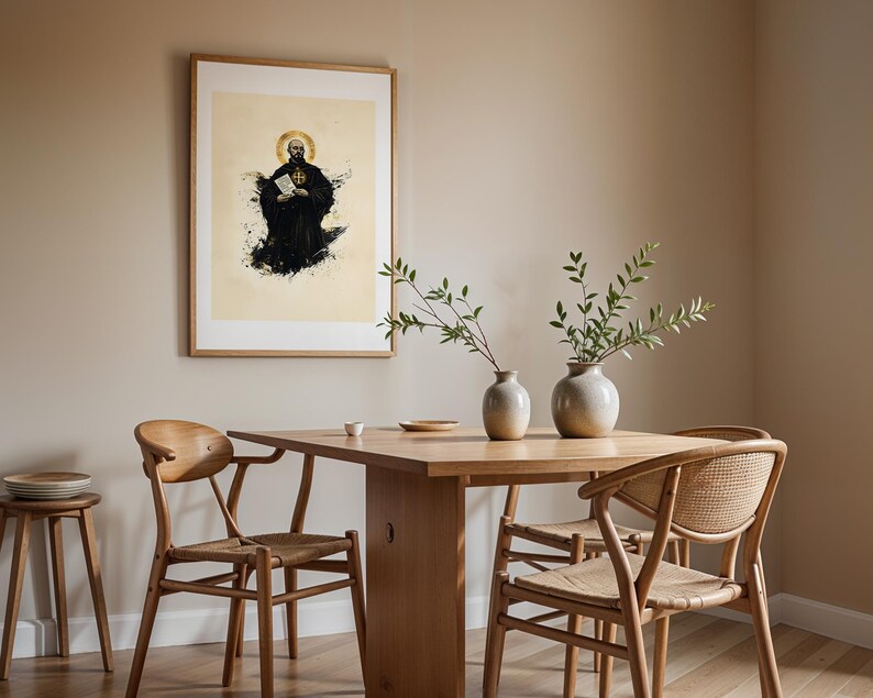 May include: A framed wall art print featuring a monk in a black habit with a golden halo, holding a book, displayed above a light wood dining table with two ceramic vases, three wooden chairs, and a small wooden stool with stacked plates.