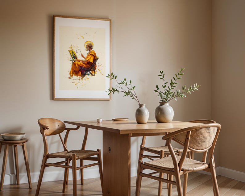 Puede incluir: Una l&aacute;mina de arte mural enmarcada que representa a una figura con t&uacute;nica naranja pintando sobre un lienzo, expuesta sobre una mesa de comedor de madera clara con dos sillas de madera clara con asientos trenzados y dos jarrones.