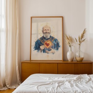 May include: A framed vertical wall art print featuring a watercolor-style portrait of a bearded man in blue vestments with a glowing, fiery heart at the center of his chest and a golden halo surrounding his head, displayed in a wooden frame.