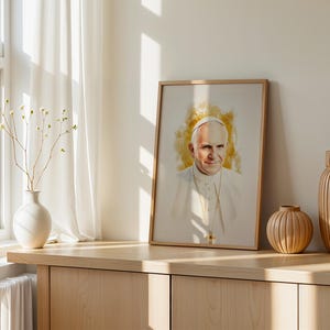 May include: A framed portrait print featuring a watercolour-style illustration of Pope John Paul II wearing a white cassock and a gold pectoral cross, set against a soft golden yellow background with a light wooden frame border.