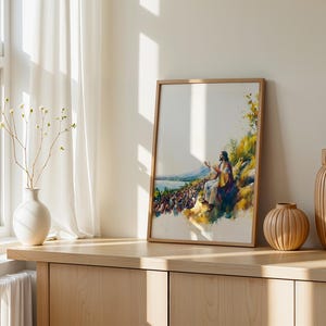 May include: A framed watercolour-style print depicting a figure in robes seated on a hillside, gesturing towards a large crowd gathered below near a body of water. The artwork features vibrant yellow, blue, and green tones with a white background.