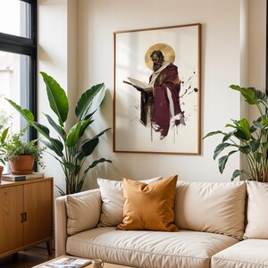 May include: A framed wall art print featuring a bearded figure in a deep burgundy robe with a gold halo, reading from an open book. The artwork has a watercolour style with paint splatters and is displayed in a thin wooden frame on a plain wall.