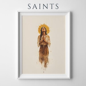 May include: A framed wall art print titled SAINTS featuring a watercolour-style illustration of a person with eyes closed and hands pressed together in prayer. The figure wears a light-coloured tunic and a cross necklace against a golden halo.
