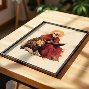 K&ouml;nnte beinhalten: Ein gerahmter Kunstdruck, der eine b&auml;rtige Gestalt in einem roten Gewand zeigt, die mit einem L&ouml;wen sitzt und in ein offenes Buch schreibt. Die Figur hat einen goldenen Heiligenschein hinter dem Kopf. Das Bild ist im Aquarellstil auf cremefarbenem Grund.