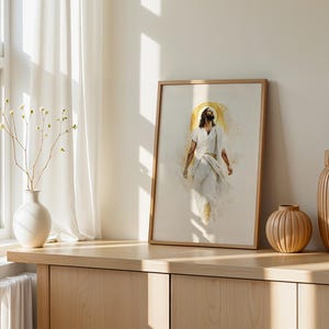 Puede incluir: Una impresi&oacute;n de arte mural vertical enmarcada que presenta una ilustraci&oacute;n estilo acuarela de una figura con t&uacute;nicas blancas y un halo dorado. La obra est&aacute; en un marco de madera clara, sobre una superficie de madera junto a un jarr&oacute;n blanco.