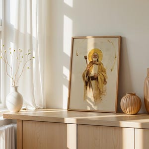 May include: A framed wall art print featuring a watercolor-style illustration of a bearded figure with a golden halo, wearing tan robes and holding a wooden staff. The artwork uses a warm, neutral color palette with soft brown and gold tones.