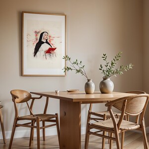 May include: A framed wall art print depicting a nun in a black and white habit holding a red book, set against a watercolor-style background. The artwork is displayed in a light wood frame on a beige wall above a wooden dining table and chairs.