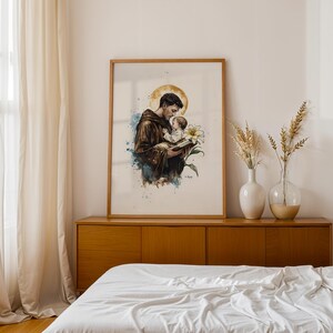Puede incluir: Una impresi&oacute;n enmarcada de estilo acuarela que representa a una figura con h&aacute;bito marr&oacute;n sosteniendo a un ni&ntilde;o peque&ntilde;o y un libro, con un halo dorado detr&aacute;s de la cabeza y una flor de lirio blanca al lado, acentuada con salpicaduras de pintura azul y marr&oacute;n.