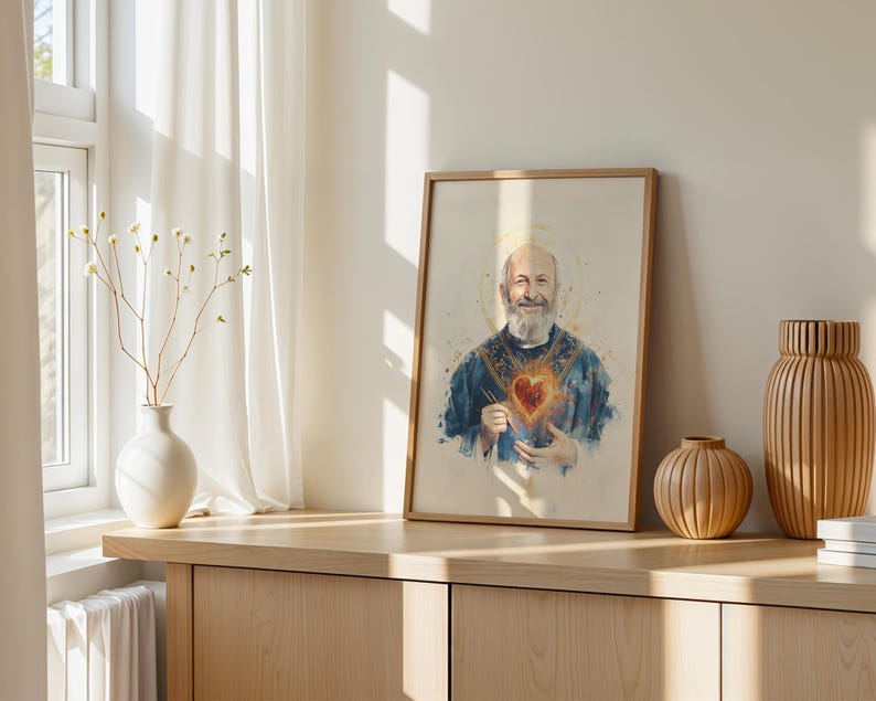 May include: A framed watercolor-style print depicting a bearded man in a blue robe with a glowing red heart on his chest and a golden halo. The artwork is displayed in a light wood frame, positioned on a wooden surface next to two vases.