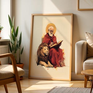 K&ouml;nnte beinhalten: Ein gerahmter Kunstdruck mit einer Aquarell-Illustration einer b&auml;rtigen Figur in rotem Gewand mit goldenem Heiligenschein, die sitzt und in ein offenes Buch schreibt, w&auml;hrend ein L&ouml;we neben ihr auf hellem cremefarbenem Hintergrund ruht.