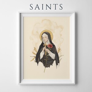 Op de afbeelding: Een ingelijste kunstdruk getiteld SAINTS met een illustratie van een figuur in een zwart habijt en witte sluier, die een houten kruis en een rode roos vasthoudt. De figuur heeft een gouden halo tegen een cr&egrave;mekleurige achtergrond met korenaren.