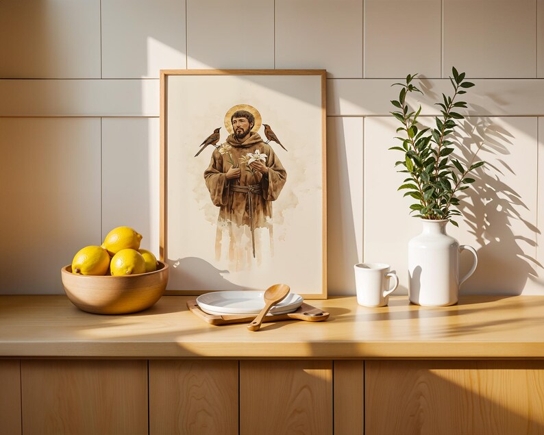 May include: A framed vertical art print depicting a bearded figure in a brown robe with a golden halo, holding white flowers with birds perched on the shoulders. The print is displayed on a wooden counter next to a bowl of yellow lemons.