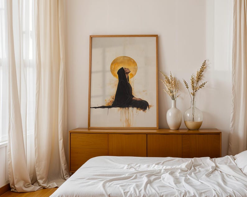 May include: A framed wall art print featuring a figure in a black hooded robe sitting in profile against a large, glowing golden circular sun. The artwork displays watercolour-style drips in shades of gold and brown on a light cream background.