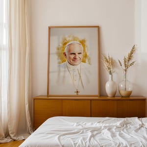 Pu&ograve; includere: Un ritratto stampato e incorniciato di Papa Giovanni Paolo II con un effetto alone dorato in stile acquerello dietro la testa. Il soggetto indossa una talare bianca e una croce pettorale dorata. L'opera &egrave; presentata in una cornice di legno chiaro.