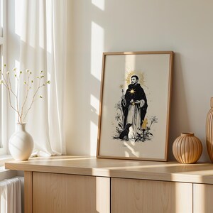 May include: A framed vertical print featuring a black and white illustration of a robed figure with a gold star on their chest, holding a flower, accompanied by a dog and floral elements, set against a light cream background with gold accents.