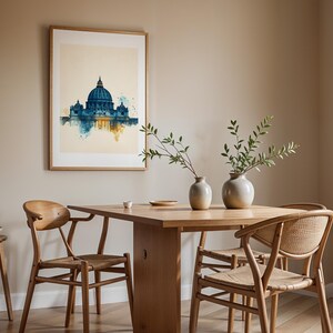 May include: A framed watercolour-style print of St. Peter's Basilica in blue and gold tones hangs on a beige wall above a wooden dining table. The table features two ceramic vases with green branches, surrounded by four wooden chairs with woven seats.