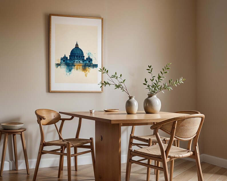 May include: A framed watercolour-style print of St. Peter's Basilica in blue and gold tones hangs on a beige wall above a wooden dining table. The table features two ceramic vases with green branches, surrounded by four wooden chairs with woven seats.