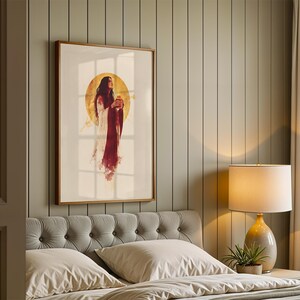 Puede incluir: Una impresi&oacute;n de arte mural vertical enmarcada que muestra una figura con cabello largo y oscuro que viste una prenda blanca y roja profunda, frente a un gran halo circular dorado. La obra se exhibe en un marco de madera fino sobre una pared panelada.