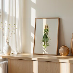 Op de afbeelding: Een ingelijste kunstdruk met een aquarelillustratie van een persoon met lang haar in een groene jurk die een zwaard vasthoudt. De figuur is omringd door delicaat groen en een vage gouden halo, gepresenteerd in een eenvoudige houten lijst.