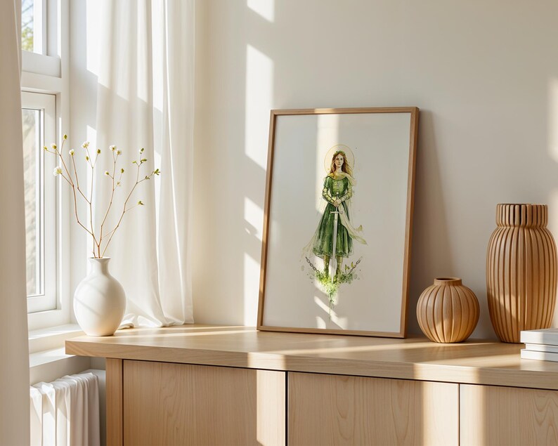 Op de afbeelding: Een ingelijste kunstdruk met een aquarelillustratie van een persoon met lang haar in een groene jurk die een zwaard vasthoudt. De figuur is omringd door delicaat groen en een vage gouden halo, gepresenteerd in een eenvoudige houten lijst.