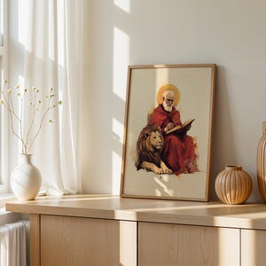Peut inclure: Une impression verticale encadr&eacute;e repr&eacute;sentant un personnage barbu en robe rouge avec une aur&eacute;ole dor&eacute;e, &eacute;crivant dans un livre tout en &eacute;tant assis &agrave; c&ocirc;t&eacute; d'un grand lion. L'&oelig;uvre est pos&eacute;e sur une surface en bois avec un vase blanc et deux vases en bois.