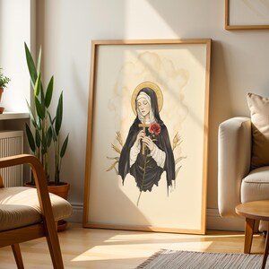 Puede incluir: Una l&aacute;mina de arte vertical enmarcada con una ilustraci&oacute;n estilizada de una monja con h&aacute;bito negro y toca blanca. Tiene un halo dorado, sostiene una cruz de madera y una rosa roja, y est&aacute; rodeada de tallos de trigo sobre un fondo crema.