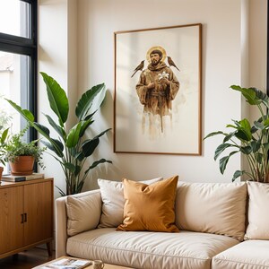 May include: A framed wall art print depicting a figure in a brown hooded robe with a golden halo, holding a white flower, with a bird perched on each shoulder. The artwork features a soft, watercolour-style aesthetic against a light background.
