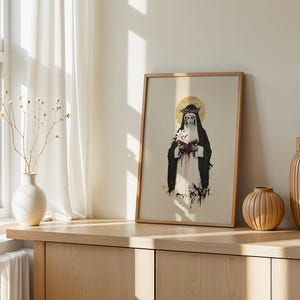 May include: A framed art print featuring a figure in a black and white habit wearing a crown of thorns with a gold halo, holding a white lily and a red book. The illustration uses ink-style lines and splashes of red, black, and gold colors.