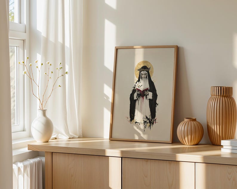 May include: A framed art print featuring a figure in a black and white habit wearing a crown of thorns with a gold halo, holding a white lily and a red book. The illustration uses ink-style lines and splashes of red, black, and gold colors.