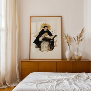May include: A framed wall art print featuring a watercolour-style illustration of a religious figure in a black and white habit, holding an open book with a golden sunburst halo behind the head, accented by brown ink splatters on a white background.