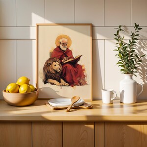 K&ouml;nnte beinhalten: Ein gerahmter Kunstdruck, der eine b&auml;rtige Figur in einem roten Gewand mit goldenem Heiligenschein zeigt, die in einem Buch schreibt und neben einem L&ouml;wen sitzt. Das Bild steht auf einer Holztheke neben einer Schale mit gelben Zitronen und einer wei&szlig;en Vase.