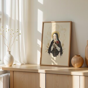 Op de afbeelding: Een ingelijste kunstdruk met een illustratie van een figuur in een zwart habijt met een gouden aureool, die een houten kruis en een rode roos vasthoudt. Het kunstwerk zit in een lichte houten lijst op een houten oppervlak naast twee vazen.