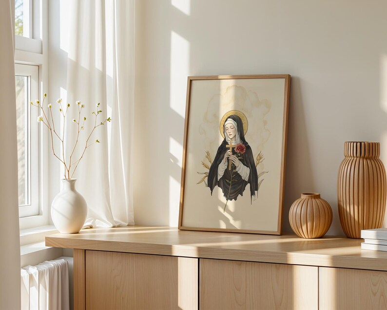 Op de afbeelding: Een ingelijste kunstdruk met een illustratie van een figuur in een zwart habijt met een gouden aureool, die een houten kruis en een rode roos vasthoudt. Het kunstwerk zit in een lichte houten lijst op een houten oppervlak naast twee vazen.