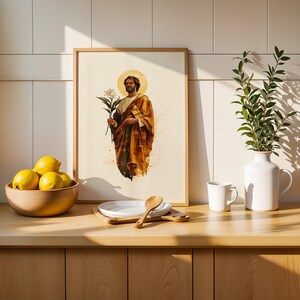 K&ouml;nnte beinhalten: Ein gerahmter Druck, der eine b&auml;rtige Figur in einem goldenen Gewand mit einer wei&szlig;en Lilie zeigt, neben einer Holzschale mit gelben Zitronen, einem Stapel wei&szlig;er Teller mit einem Holzl&ouml;ffel, einer wei&szlig;en Tasse und einem wei&szlig;en Krug mit Gr&uuml;n.
