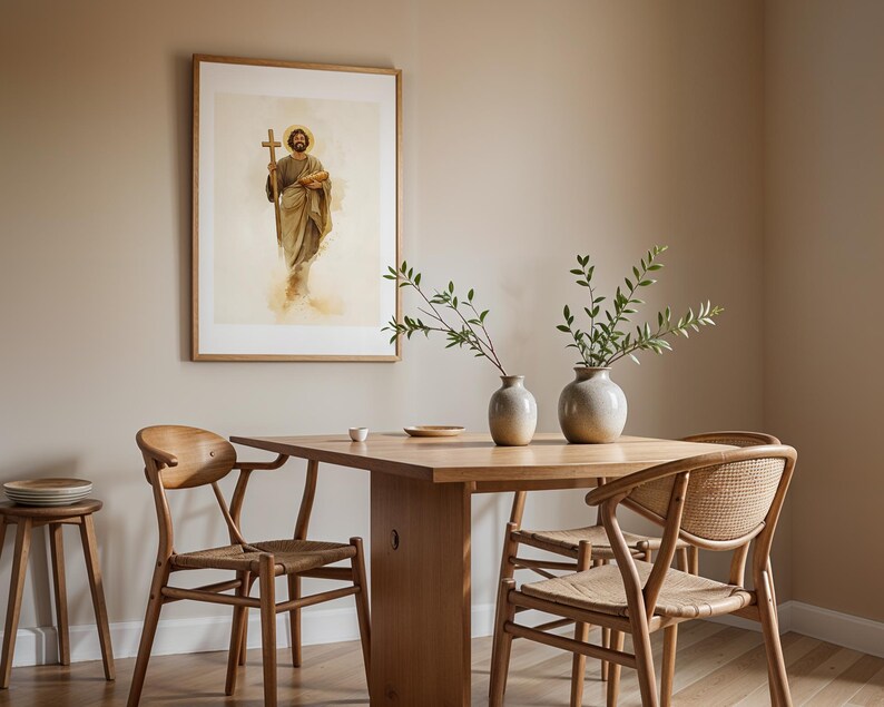 Op de afbeelding: Een ingelijste kunstdruk met een figuur die een houten kruis en een brood vasthoudt, boven een houten eettafel met twee stoelen en twee keramische vazen met groene bladeren, naast een kleine houten kruk.