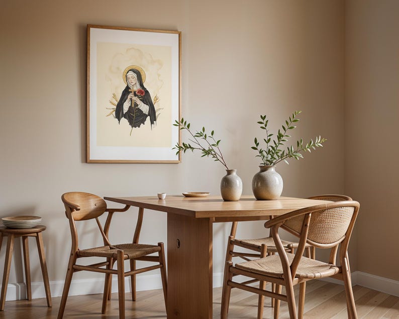 Op de afbeelding: Een ingelijste muurkunstprint met een figuur in een zwart habijt met een gouden aureool, die een houten kruis en een rode roos vasthoudt. De print hangt in een kamer met een houten eettafel en vier houten stoelen met gevlochten zittingen.