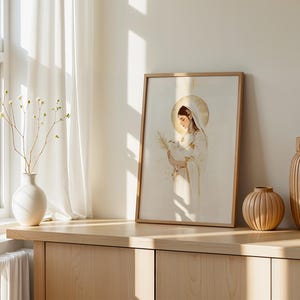 Pode incluir: Uma impress&atilde;o de arte emoldurada com uma ilustra&ccedil;&atilde;o de linhas de uma figura com um halo dourado, segurando um pequeno p&aacute;ssaro e uma folha de palmeira. A obra usa tons suaves de dourado e branco, apresentada numa moldura de madeira clara.