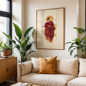 May include: A framed wall art print featuring a figure in a deep red robe with a golden halo, set against a cream background. The artwork displays a stylised, painterly aesthetic with scattered red accents surrounding the central figure.