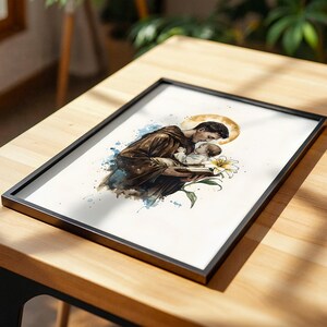 Puede incluir: Una l&aacute;mina art&iacute;stica enmarcada con una ilustraci&oacute;n en acuarela de una persona con t&uacute;nica marr&oacute;n que sostiene a un ni&ntilde;o peque&ntilde;o. Un halo dorado brilla detr&aacute;s de la cabeza de la persona y una flor de lirio blanco se sit&uacute;a en primer plano con salpicaduras.