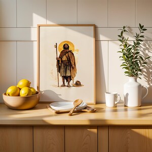 May include: A framed wall art print featuring a watercolour illustration of a bearded figure in a brown cloak holding a wooden staff, standing before a yellow halo. The artwork is displayed on a wooden counter next to a bowl of lemons and a vase.
