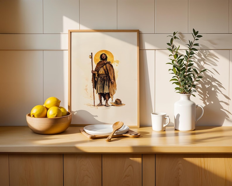 May include: A framed wall art print featuring a watercolour illustration of a bearded figure in a brown cloak holding a wooden staff, standing before a yellow halo. The artwork is displayed on a wooden counter next to a bowl of lemons and a vase.