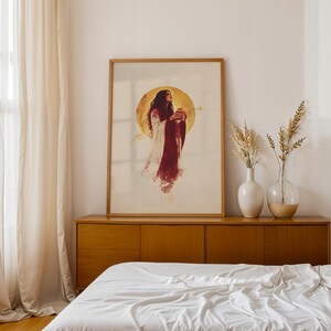 Puede incluir: Una impresi&oacute;n de arte mural vertical enmarcada que muestra una figura con cabello largo y oscuro vistiendo una prenda fluida blanca y roja oscura, frente a un gran halo circular dorado. La obra est&aacute; en un marco de madera clara.