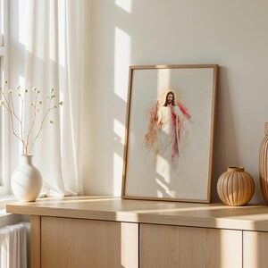 May include: A framed wall art print featuring a watercolor-style illustration of a figure with long hair and a beard, wearing white robes with red accents, surrounded by a golden halo, displayed in a light wood frame on a wooden surface.