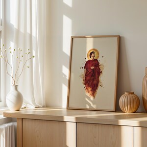 May include: A framed vertical art print featuring a watercolor-style illustration of a figure in a deep red robe with a golden halo. The artwork is displayed in a light-toned wooden frame, positioned on a wooden surface alongside two vases.