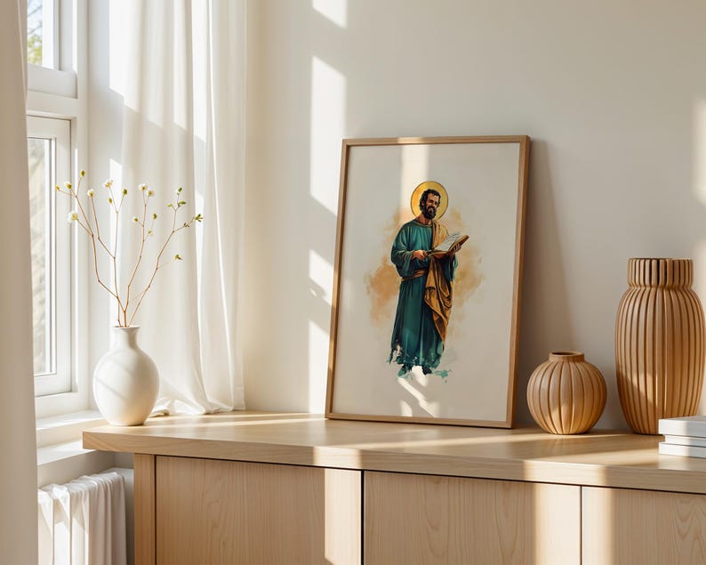 Op de afbeelding: Een ingelijste verticale print met een aquarelillustratie van een bebaard figuur in een blauwgroen gewaad en gouden mantel die een open boek vasthoudt, met een gouden halo boven het hoofd, in een lichthouten lijst op een houten oppervlak.