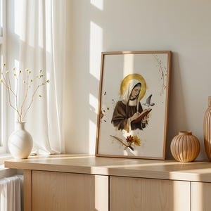 May include: A framed wall art print featuring a figure in a brown habit and white head covering writing in a book, with a golden halo, a flying bird, and floral accents. The illustration is rendered in a soft, artistic style on a cream background.