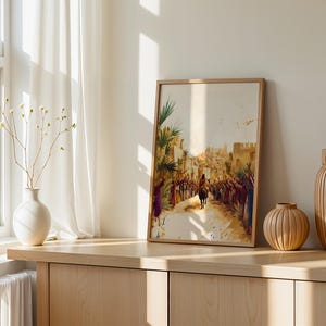May include: A framed watercolour-style print depicting a figure riding a donkey through a crowd in a desert town, featuring warm earth tones, palm trees, and stone architecture. The artwork is displayed in a light wood frame on a wooden surface.