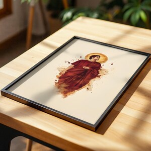 May include: A framed rectangular print featuring a watercolor-style illustration of a figure in a deep red robe with a golden halo behind their head. The artwork is set against a cream-colored background with scattered brown and red paint splatters.