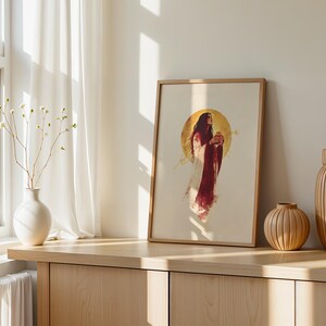 Puede incluir: Una impresi&oacute;n de arte mural vertical enmarcada que muestra una figura con cabello largo y oscuro que viste una prenda fluida de color blanco y rojo intenso, colocada sobre un gran fondo circular dorado. La obra est&aacute; en un marco de madera clara.